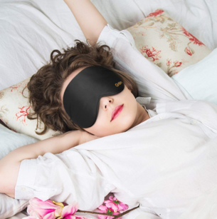 Une femme s'allonge confortablement sur un oreiller fleuri dans son lit, les bras étirés après une bonne nuit, avec un masque de sommeil Gritin noir (Page de cluster 1 - Topic Cluster)